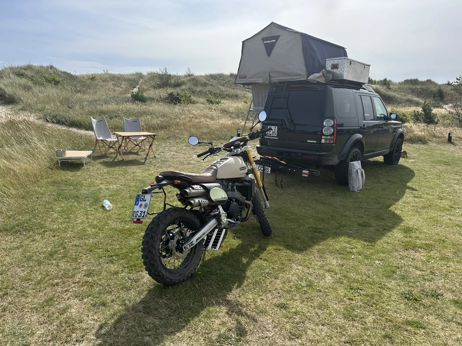 Land Rover mit geöffnetem Dachzelt und Motorrad auf einem Stellplatz zwischen Dünen